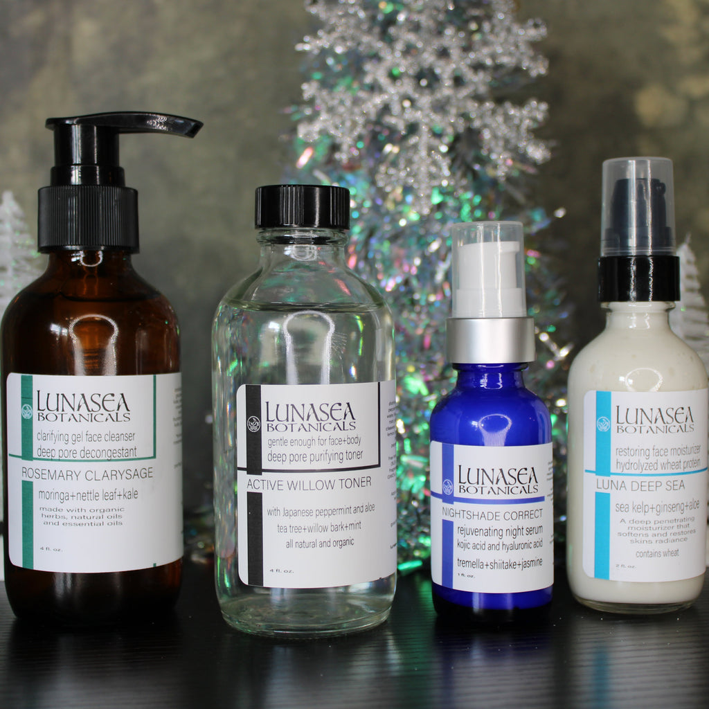 Four bottles of Lunasca Botanicals skincare products on a dark surface with a decorative Christmas tree in the background.