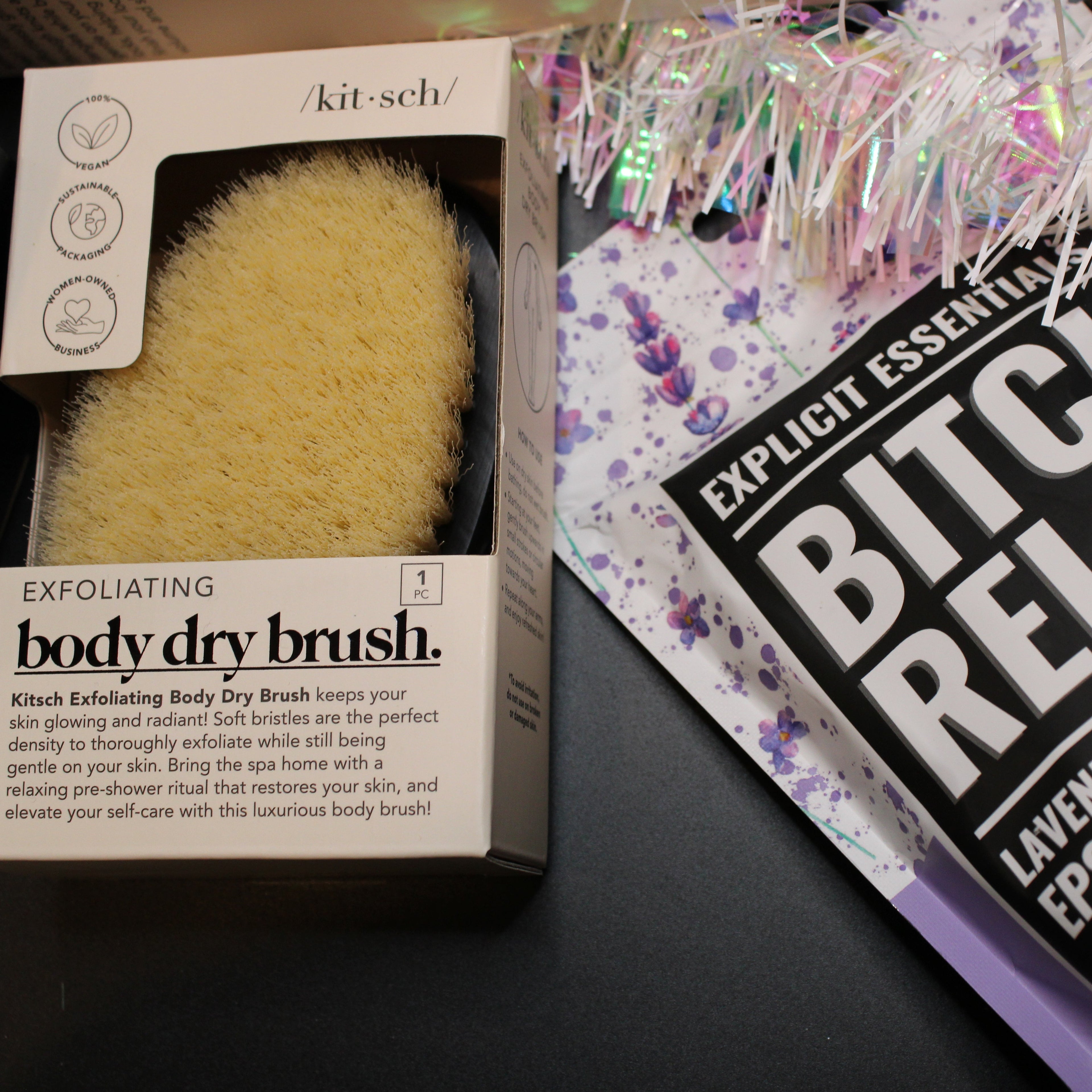 Body dry brush and bath salts packaging on a dark surface with decorative tinsel.