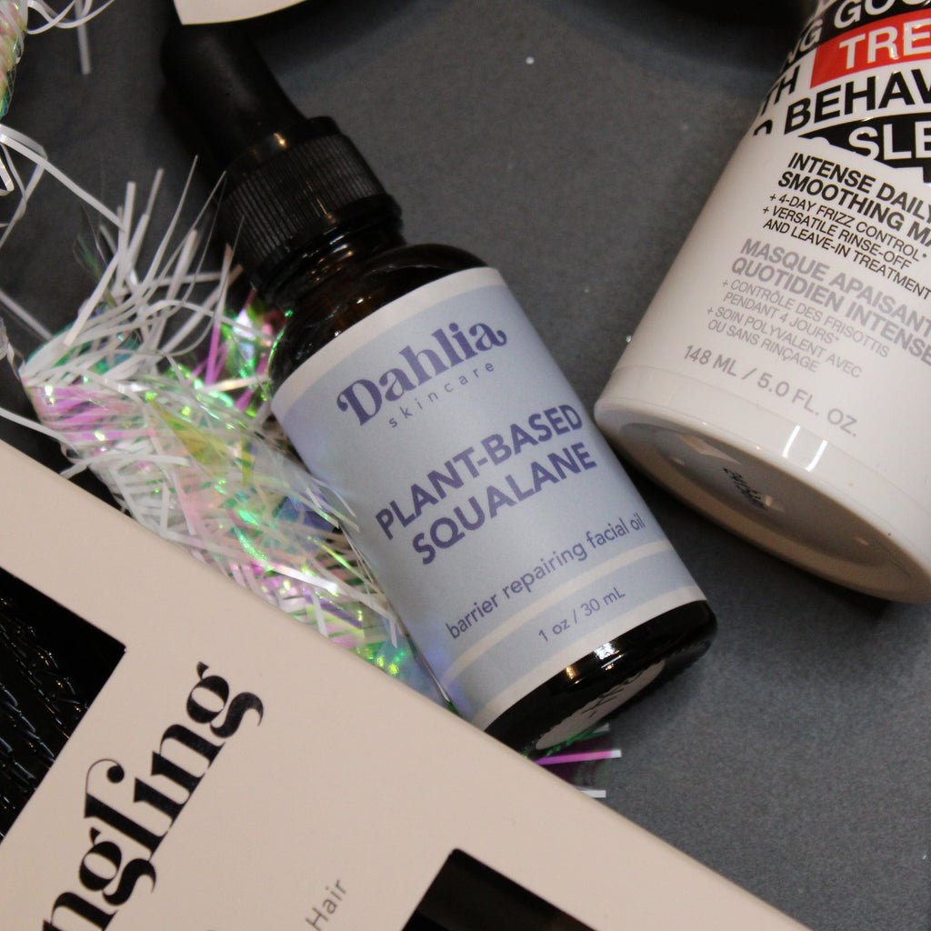 Cosmetic products including a bottle of 'Dahlila' and a container labeled 'Good Behavior' on a dark surface.