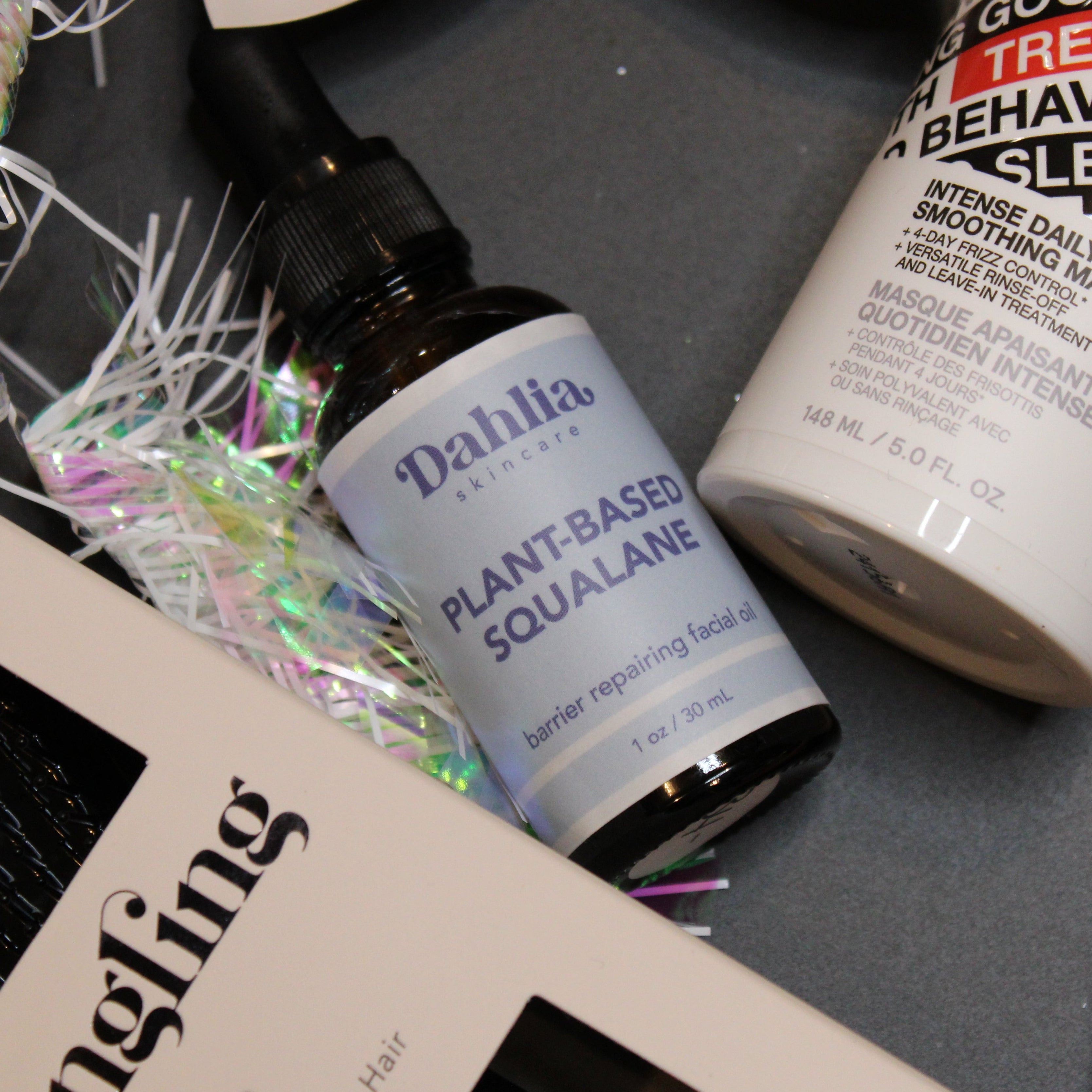 Cosmetic products including a bottle of 'Dahlila' and a container labeled 'Good Behavior' on a dark surface.