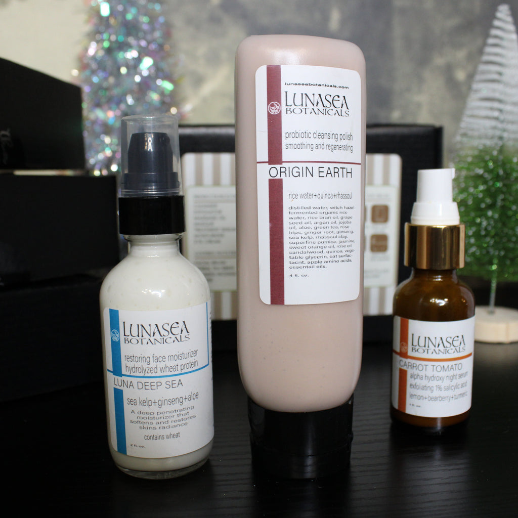 Three skincare products from Elinasa Botanicals on a dark surface with decorative items in the background.